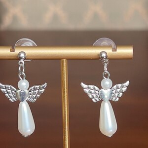 May include: A pair of angel-shaped earrings. Each earring features a silver-colored wing design, a heart-shaped center, a white pearl bead, and a teardrop-shaped pearl pendant. The earrings are displayed on a gold-colored stand.