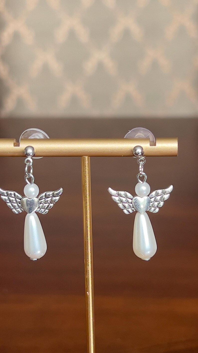 May include: A pair of angel-shaped earrings. Each earring features a silver-colored wing design, a heart-shaped center, a white pearl bead, and a teardrop-shaped pearl pendant. The earrings are displayed on a gold-colored stand.