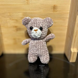 Könnte beinhalten: Ein kleiner, handgehäkelter Teddybär in einem weichen, gedämpften Lila. Der Bär hat schwarze Knopfaugen, eine weiße Schnauze und eine hellbraune Nase. Seine Arme sind ausgestreckt, und er steht auf zwei kleinen Beinen. Ein bezaubernder, handgefertigter Artikel.