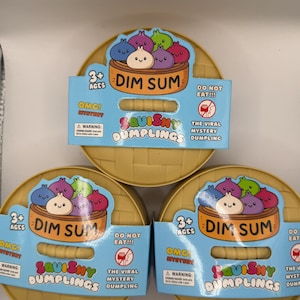 May include: Three boxes of Squishy Dumplings, a mystery toy. Each box has a yellow, round container with a blue label featuring a cartoon dim sum design. The text on the label includes "Dim Sum", "Squishy Dumplings", and "Do Not Eat!!!".