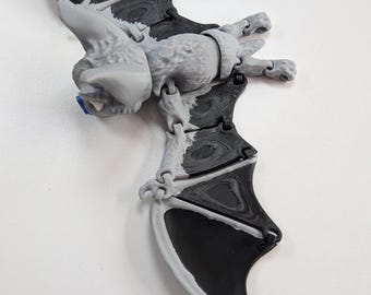 May include: An articulated bat figurine featuring a light gray body and head with contrasting black wing membranes. The figure has a textured surface, small ears, and a blue accent on the nose area, with multiple joints visible along the wings.