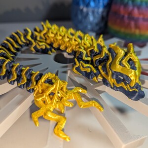 May include: A 3D-printed articulated dragon figurine in navy blue and gold. The dragon is coiled and rests on a white geometric base. The dragon's body is navy blue with gold accents, and the head and tail are also gold.