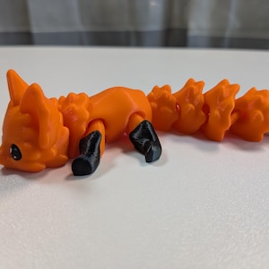 Könnte beinhalten: Eine bewegliche Fuchsfigur mit einem leuchtend orangefarbenen Körper, schwarzen Pfoten und einer weiß gespitzten Rute. Der Fuchs hat ein segmentiertes, flexibles Design mit sichtbaren 3D-Druckschichten, spitzen Ohren und kleinen schwarzen Augen.