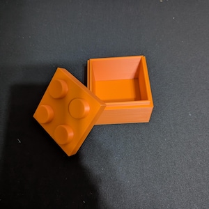May include: A square orange storage box with a removable lid. The lid features four raised circular studs on its top surface, mimicking the appearance of a building block. The box is open, revealing a hollow interior and straight walls.