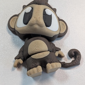 May include: A 3D printed monkey figurine featuring a dark brown and tan color scheme. The character has large white eyes with black pupils, prominent ears, a curled tail, and visible layer lines throughout the textured plastic surface.