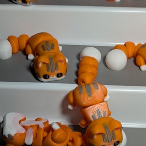 Op de afbeelding: Oranje en witte kattenbeeldjes met zwarte details zijn gerangschikt op een wit en grijs oppervlak. De kattenbeeldjes hebben een gesegmenteerd lichaam en een witte bal aan het einde van de staart. De katten staan in een speelse pose.