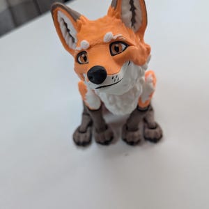 May include: A 3D printed fox figurine featuring a bright orange coat, white chest and facial markings, and dark brown paws and ear tips. The fox has large, expressive eyes, a black nose, and a textured surface characteristic of additive manufacturing.