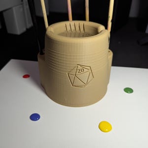 May include: A tan-colored, cylindrical object with a geometric design and the number "20" printed on it. The object is surrounded by thin, vertical rods. Colorful paint dots in red, green, blue, and yellow are scattered on the white surface.