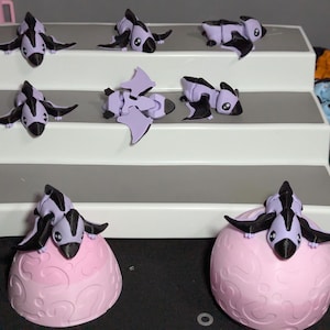 May include: A selection of small, purple and black toy creatures with outstretched wings and beaks, displayed on a tiered stand. Two of the creatures are on pink, patterned egg-shaped bases. The background includes other toys and a black surface.