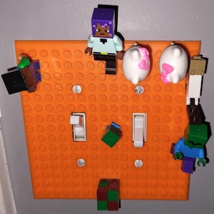 May include: An orange Lego-style light switch plate with two light switches.  There are several small Lego minifigures and blocks attached to the plate, including a purple-haired minifigure, two white pigs, a green block, and a blue zombie minifigure.