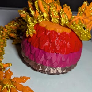 May include: A detailed, multi-colored dragon figurine. The dragon features a gold head and spine, with an orange and red body. The base is a layered sphere of red, pink, and brown. A matching orange flower is in the foreground.