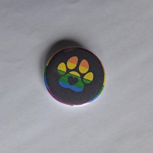 May include: A circular badge featuring a rainbow paw print on a black background, with a heart in the centre of the paw. The edge of the badge has a rainbow gradient.