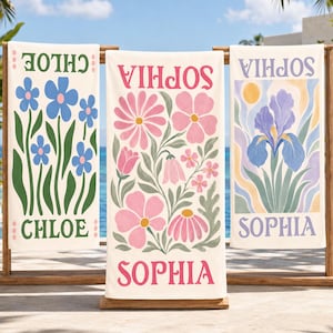 May include: Three rectangular beach towels hanging on a wooden rack. The left towel features blue flowers and the name CHLOE in green. The center towel displays pink flowers and the name SOPHIA in pink. The right towel shows a purple iris and SOPHIA.