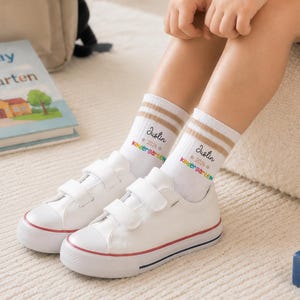 May include: White crew socks with two tan stripes at the top, featuring the printed text "Justin", a star icon, and the word "kindergarten" in rainbow-colored letters. The socks are worn with white low-top sneakers that have two hook-and-loop straps.