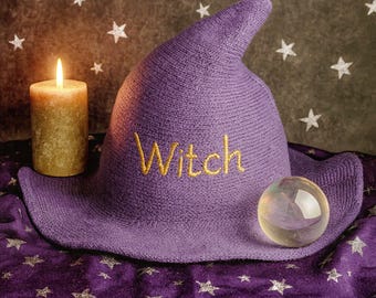 Puede incluir: Un sombrero de bruja morado con la palabra "Witch" bordada en dorado. Una vela encendida y una bola de cristal están colocadas junto al sombrero sobre una tela morada con patrones de estrellas. El fondo es marrón oscuro con estrellas blancas.