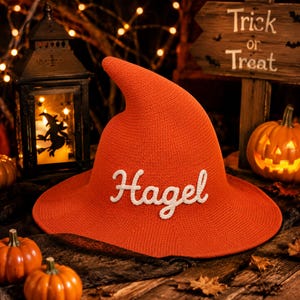 May include: A bright orange knitted witch hat featuring the word Hagel in white cursive lettering on the front. The hat has a pointed crown and a wide brim, constructed from a textured yarn material with a consistent stitch pattern throughout.