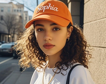 May include: Orange baseball cap with the name "Sophia" embroidered in white script. The cap is worn by a person wearing a white t-shirt and gold chain necklace. The background shows a city street.