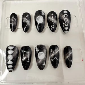 May include: A set of ten black press-on nails with celestial designs. Each nail features a glossy black base with white and silver accents, including stars, constellations, and moon phases. The nails are oval-shaped and displayed in a clear plastic case.