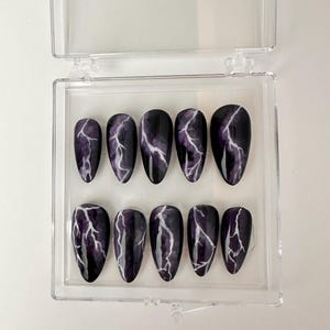 May include: A set of ten false nails in a clear plastic case. The nails are a dark purple color with a white lightning bolt design.
