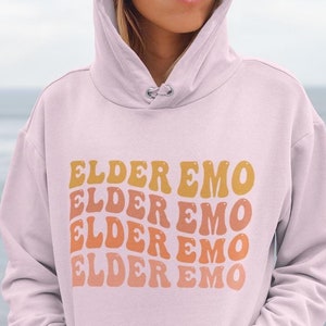 May include: A light pink hoodie with the text "ELDER EMO" repeated four times in a retro font. The text is in shades of orange and yellow.