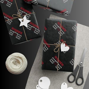 Könnte beinhalten: Mehrere Geschenkverpackungen mit schwarzem Papier, bedruckt mit weißen Häusern und dem Wort "MAYHEM" in Rot. Die Geschenke sind mit Bindfaden verschnürt und haben weiße stern- und herzförmige Anhänger. Eine Spule weißen Bindfadens und eine Schere sind ebenfalls vorhanden.