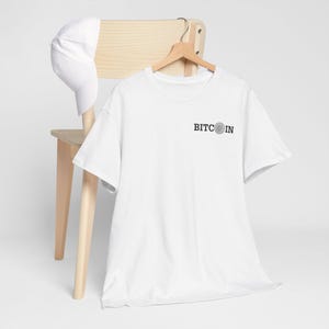 May include: A white t-shirt with the word "BITCOIN" printed in black. The shirt is hanging on a wooden hanger. A white baseball cap rests on the back of a wooden chair.