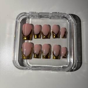 May include: A set of ten pink and gold press-on nails in a clear plastic case. The nails have a light pink base with a gold metallic French tip design. The nails are oval-shaped and vary in size.