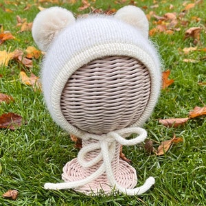 May include: White knitted baby hat with fluffy pom poms on top and a white tie. The hat is on a pink wicker stand.