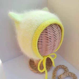 May include: A light green and yellow knitted baby bonnet with fuzzy yarn and a yellow tie. The bonnet is on a wicker hat form.