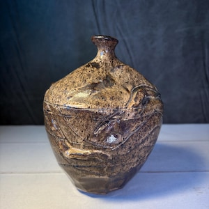 Może przedstawiać: Ręcznie wykonany, teksturowany ceramiczny słoik z pokrywką. Słoik jest głównie brązowy z jaśniejszymi beżowymi akcentami i ciemnobrązową podstawą. Naczynie ma pękaty korpus i wąską szyjkę.
