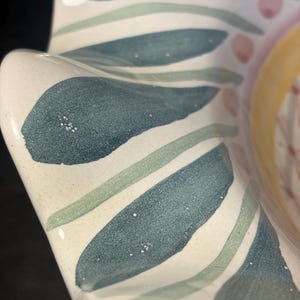Op de afbeelding: Een close-up van een keramische kom met een witte basis en een bloemenontwerp. Het ontwerp bevat blauwe, groene en roze geschilderde bladeren en accenten. De kom heeft een golvende, geschulpte rand.