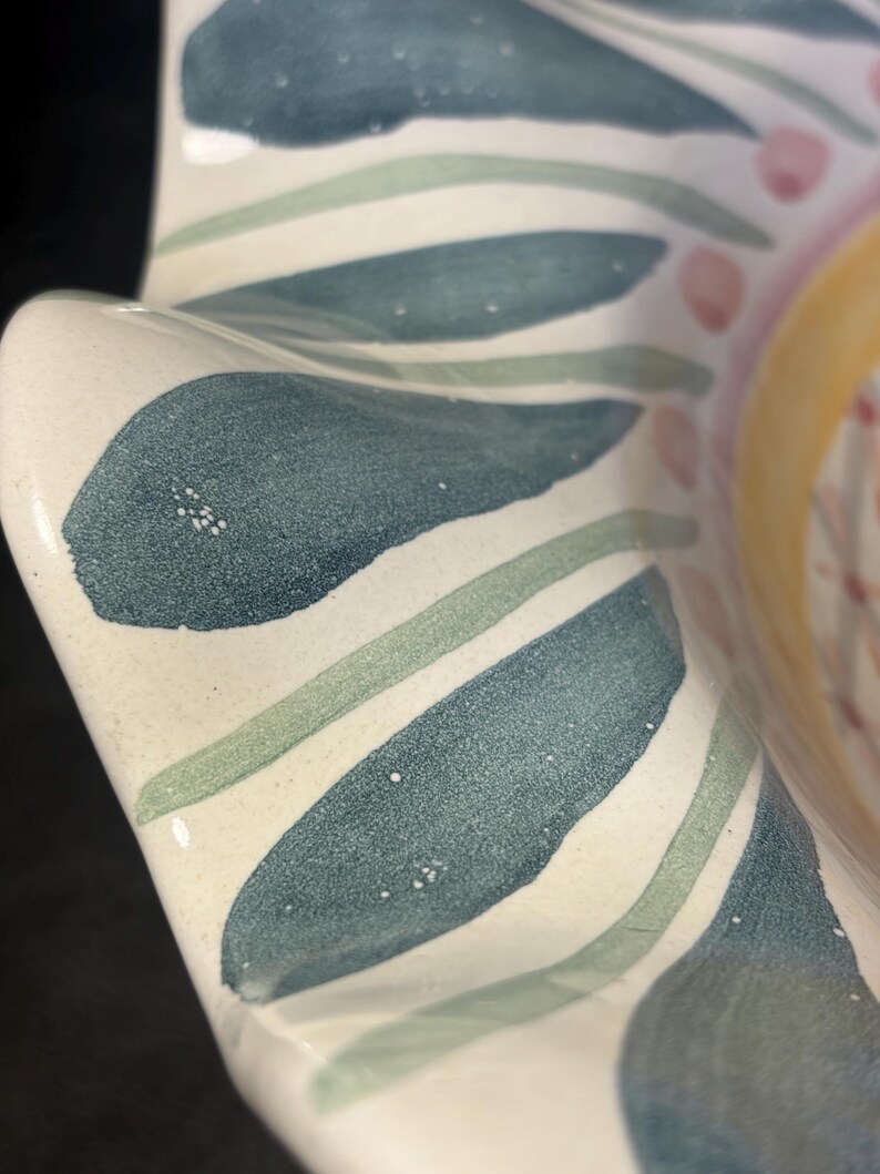 Op de afbeelding: Een close-up van een keramische kom met een witte basis en een bloemenontwerp. Het ontwerp bevat blauwe, groene en roze geschilderde bladeren en accenten. De kom heeft een golvende, geschulpte rand.