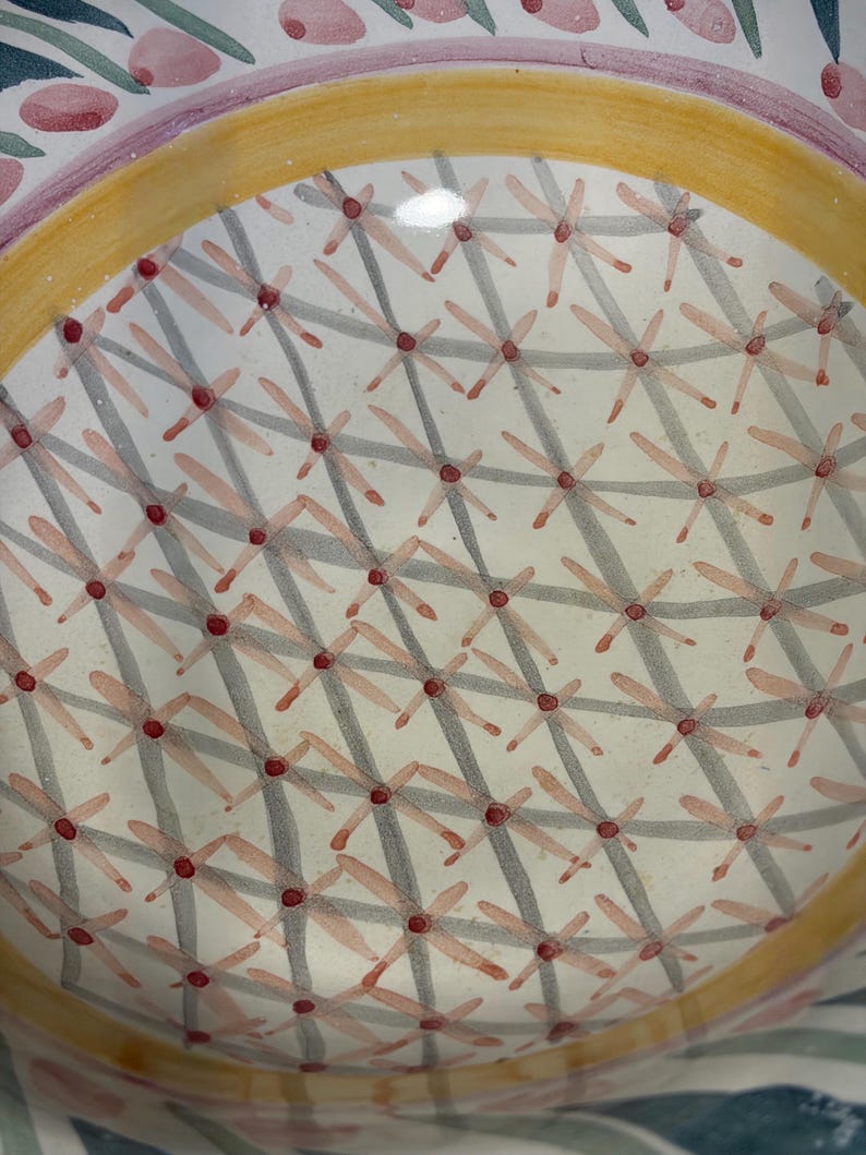 Op de afbeelding: Close-up van een decoratieve keramische plaat met een bloemenontwerp. De plaat heeft een witte achtergrond met een herhalend patroon van grijze kruisarcering en roze bloemachtige vormen. Een gele band omcirkelt het midden en de rand heeft roze en groene bloemaccenten.