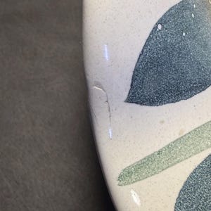 Op de afbeelding: Close-up van een keramisch object met een wit gespikkeld oppervlak. Het ontwerp bevat teal en groene bladachtige patronen. Een kleine chip is zichtbaar op de rand van het object.