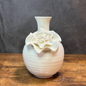 May include: Cream-colored ceramic vase with a large, sculpted flower detail. The vase has a rounded base with horizontal ridges and a slender neck. The flower is positioned around the vase's midsection, adding a decorative touch to the floral design.