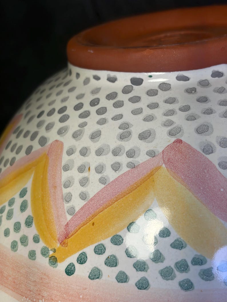 Op de afbeelding: Een keramische kom met een witte basis, versierd met een patroon van grijze stippen en kleurrijke geometrische ontwerpen. De kom heeft roze, gele en groene accenten en een bruine rand.