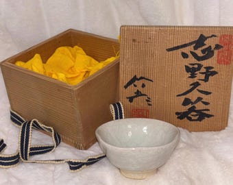 Könnte beinhalten: Eine Holzkiste mit gelber Stoffauskleidung und japanischen Schriftzeichen auf dem Deckel. Die Kiste steht neben einer kleinen, weißen Keramikschale.