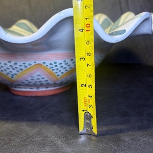 Op de afbeelding: Een keramische kom met een geschulpte rand, met een handgeschilderd ontwerp. De kom heeft een diameter van ongeveer 12.7 cm. Het ontwerp omvat blauwe en groene strepen, oranje en gele accenten en kleine blauwe stippen.