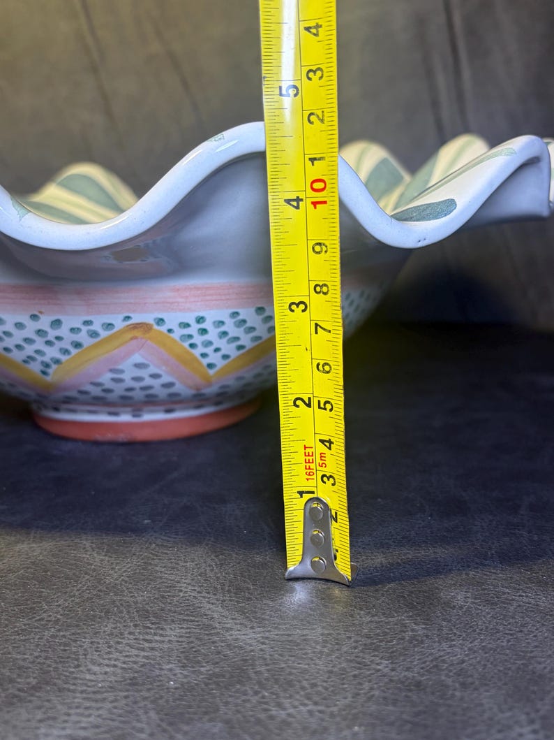 Op de afbeelding: Een keramische kom met een geschulpte rand, met een handgeschilderd ontwerp. De kom heeft een diameter van ongeveer 12.7 cm. Het ontwerp omvat blauwe en groene strepen, oranje en gele accenten en kleine blauwe stippen.