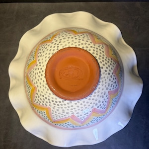 Op de afbeelding: Een keramische kom met een witte, gegolfde rand. De binnenkant van de kom heeft een wit oppervlak met een patroon van kleine grijze stippen en roze en gele zigzagontwerpen. De bodem van de kom is terracotta.