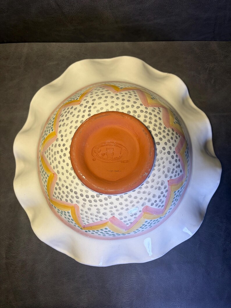 Op de afbeelding: Een keramische kom met een witte, gegolfde rand. De binnenkant van de kom heeft een wit oppervlak met een patroon van kleine grijze stippen en roze en gele zigzagontwerpen. De bodem van de kom is terracotta.