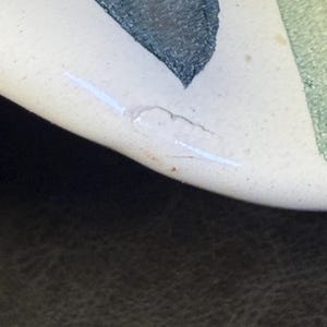 Op de afbeelding: Close-up van een keramisch stuk met een cr&egrave;mekleurig oppervlak. Het ontwerp toont abstracte vormen in tinten groen en donkerblauw. Een witte, lineaire markering en een kleine, onregelmatige witte vorm zijn zichtbaar.