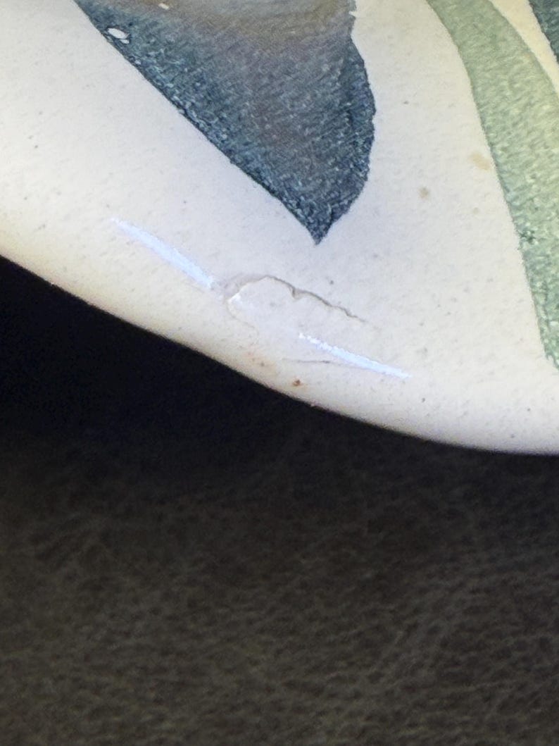 Op de afbeelding: Close-up van een keramisch stuk met een cr&egrave;mekleurig oppervlak. Het ontwerp toont abstracte vormen in tinten groen en donkerblauw. Een witte, lineaire markering en een kleine, onregelmatige witte vorm zijn zichtbaar.