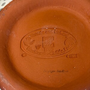 Op de afbeelding: Close-up van de bodem van een terracotta-kleurige keramische kom. De basis heeft een reli&euml;f ovaal met de tekst "Victoria and Richard", "Mackenzie-Childs, Ltd.", "1990" en het nummer "01803".