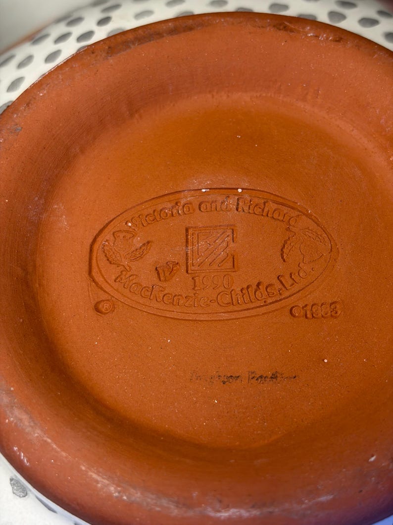Op de afbeelding: Close-up van de bodem van een terracotta-kleurige keramische kom. De basis heeft een reli&euml;f ovaal met de tekst "Victoria and Richard", "Mackenzie-Childs, Ltd.", "1990" en het nummer "01803".