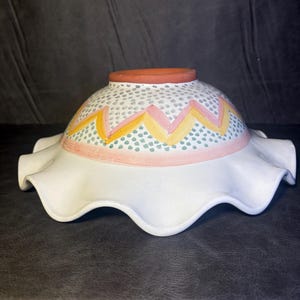 Op de afbeelding: Een witte keramische kom met een gegolfde rand. De kom heeft een decoratieve band met roze, gele en groene accenten. De bovenste rand is terracotta van kleur.