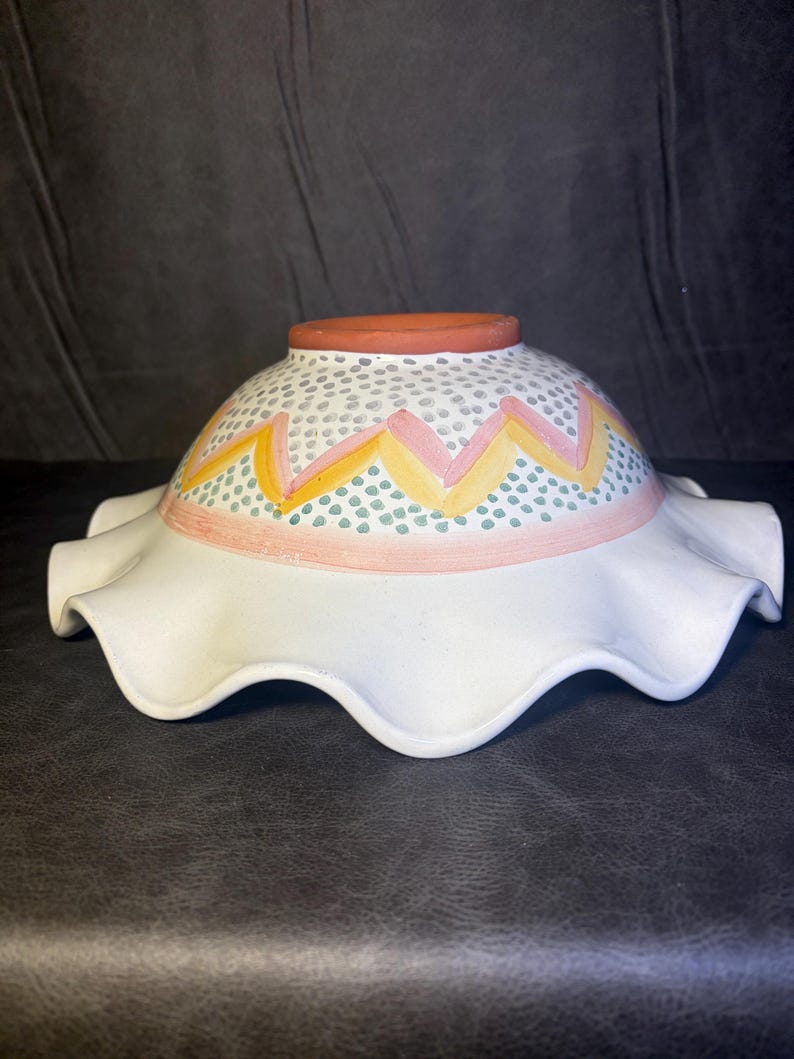 Op de afbeelding: Een witte keramische kom met een gegolfde rand. De kom heeft een decoratieve band met roze, gele en groene accenten. De bovenste rand is terracotta van kleur.