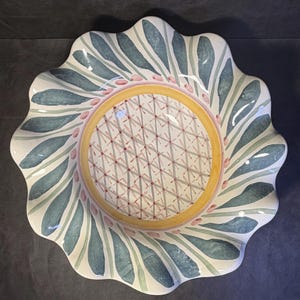 Op de afbeelding: Een decoratieve keramische schaal met een geschulpte rand. De schaal heeft een centrale gele cirkel met een geometrisch patroon, omgeven door een ring van roze stippen en blauwe en groene bladachtige ontwerpen. De schaal heeft een witte achtergrond.