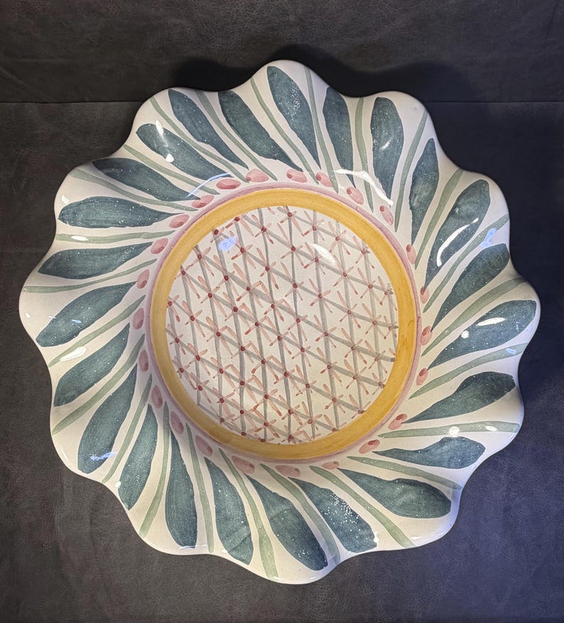 Op de afbeelding: Een decoratieve keramische schaal met een geschulpte rand. De schaal heeft een centrale gele cirkel met een geometrisch patroon, omgeven door een ring van roze stippen en blauwe en groene bladachtige ontwerpen. De schaal heeft een witte achtergrond.