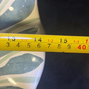 Op de afbeelding: Een gedeelte van een keramische vaas met een geschilderd bladmotief in tinten blauw en groen op een cr&egrave;mekleurige achtergrond. Een gele meetlint toont een lengte van ongeveer 35,5 cm.